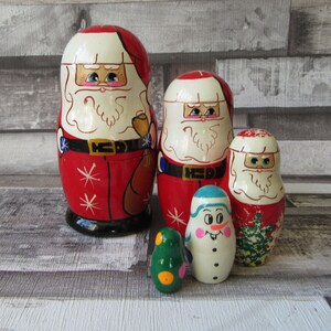 Pode incluir: Um conjunto de cinco bonecas russas, com o Pai Natal e um boneco de neve. As bonecas são pintadas em vermelho, branco e verde, com cintos pretos e fivelas douradas. A boneca maior tem cerca de 15 cm de altura.