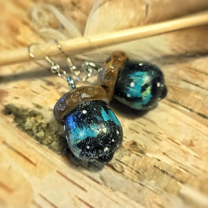 May include: A pair of handmade acorn earrings. The acorn caps are brown, and the bodies are painted with a galaxy design in shades of blue, black, and white. The earrings hang from silver-colored hooks.