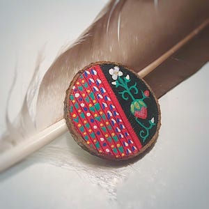May include: A small wooden circle with a red, white and blue geometric pattern on one half and a black background with a green stem and red strawberry on the other half.