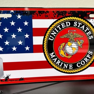 May include: A red, white and blue American flag with a black and white distressed background. The flag has two dog tags hanging from a chain. The dog tags are silver and blank. The flag is centred on the licence plate with the words "United States Marine Corps" in a gold circle.