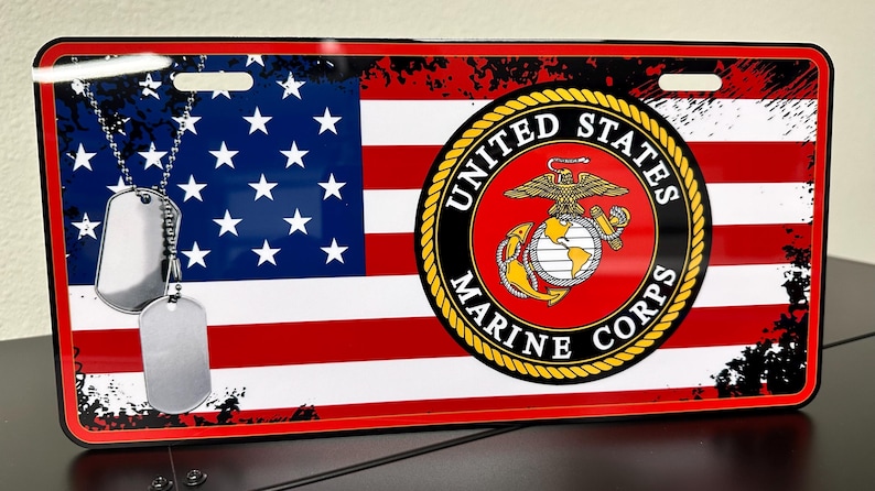May include: A red, white and blue American flag with a black and white distressed background. The flag has two dog tags hanging from a chain. The dog tags are silver and blank. The flag is centred on the licence plate with the words "United States Marine Corps" in a gold circle.