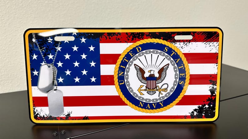 May include: A metal licence plate featuring a distressed American flag with a navy blue seal that reads "United States Navy". Two silver dog tags hang from the top of the plate.