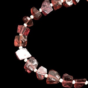 May include: A close-up of a red gemstone necklace with white beads. The necklace is made of faceted, irregular shaped stones.