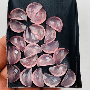 Puede incluir: Colección de gemas de cuarzo rosa facetadas, semicirculares, en una bandeja negra. Las gemas son de color rosa suave y tienen una superficie pulida y reflectante. La bandeja está sostenida por una mano.