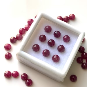 May include: A collection of polished, round, deep red gemstones, possibly rubies, displayed in a white square container and scattered on a white surface. The gems have a smooth, reflective surface, and vary slightly in color intensity.
