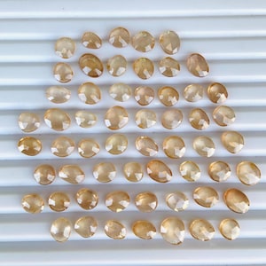 May include: A collection of 50 loose, faceted, oval-shaped, light brown gemstones. The gemstones are arranged in a semi-circle pattern on a white surface.