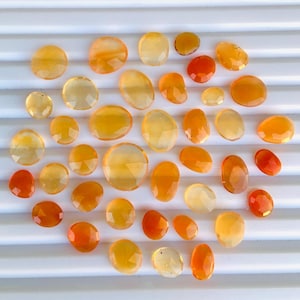 Puede incluir: Una colección de 30 cabujones de piedras preciosas de color naranja y amarillo. Los cabujones son de varias formas y tamaños, algunos son ovalados y otros redondos. Los cabujones están dispuestos en un círculo sobre una superficie blanca.