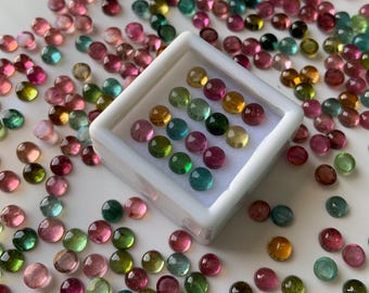 May include: A collection of round, multi-colored gemstones in a white box. The gemstones are various shades of green, pink, and brown. The gemstones are scattered around the box.