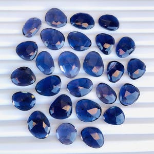 Peut inclure: Une collection de 25 pierres précieuses bleu foncé de forme ovale facettées. Les pierres sont disposées en cercle sur un fond blanc.