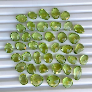 Peut inclure: Une collection de 35 pierres précieuses de péridot vert, chacune avec une forme facetée unique. Les pierres sont disposées sur une surface blanche.