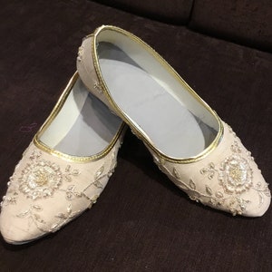 Op de afbeelding: Een paar cr&egrave;mekleurige instapschoenen met ingewikkeld goudkleurig borduurwerk in een bloemmotief. De schoenen hebben een puntige neus, een metallic gouden bies langs de rand en een lichtgekleurde binnenvoering met gedetailleerd naaiwerk.