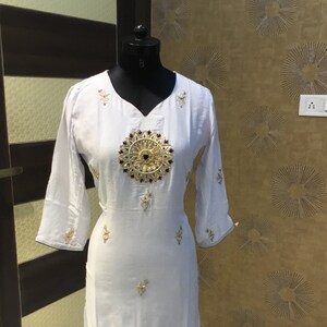 Op de afbeelding: Een witte knielange tuniek met driekwartmouwen en een V-hals. Het kledingstuk is voorzien van een groot, rond goud- en donkerblauw geborduurd medaillon op het midden van de borst, aangevuld met kleinere gouden bloemenborduursels verspreid over de stof.