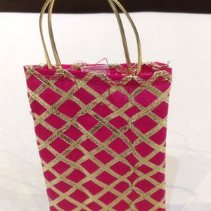 Pode incluir: Uma pequena bolsa retangular de tecido rosa choque com um padrão de treliça metálico dourado. A bolsa inclui duas alças metálicas douradas finas presas à borda superior e um pequeno anel de metal prateado visível no centro da base.