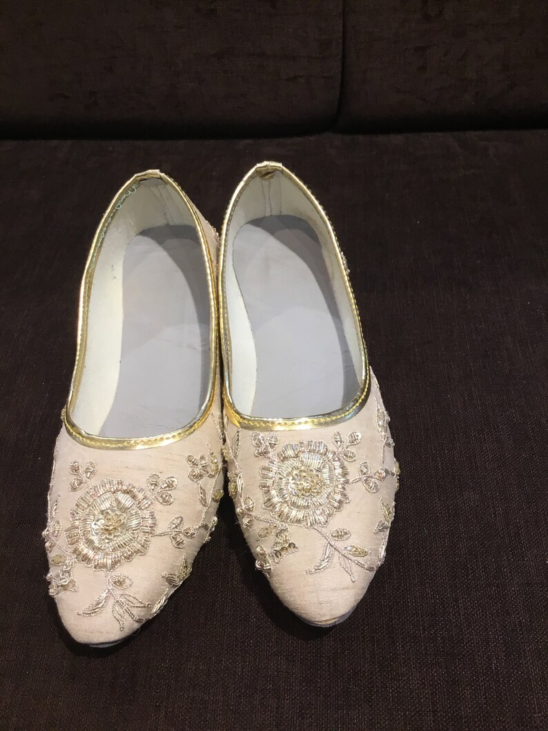 Op de afbeelding: Een paar beige instappers met een puntige neus en een goudkleurige bies langs de opening. De buitenkant is versierd met gedetailleerd goudkleurig borduurwerk in een bloemmotief, en de binnenzijde is voorzien van een lichtgrijze voering.