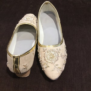 Op de afbeelding: Een paar beige instappers met gedetailleerd goudkleurig borduurwerk in een bloemmotief op de neus en zijkanten. De schoenen hebben een puntige neus, een goudkleurige bies langs de rand en een lichtgrijze voering aan de binnenzijde.