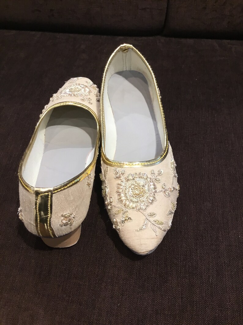 Op de afbeelding: Een paar beige instappers met gedetailleerd goudkleurig borduurwerk in een bloemmotief op de neus en zijkanten. De schoenen hebben een puntige neus, een goudkleurige bies langs de rand en een lichtgrijze voering aan de binnenzijde.