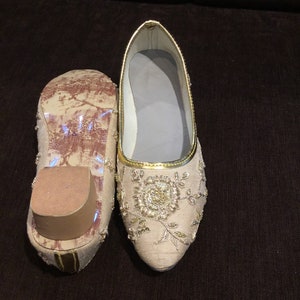 Op de afbeelding: Een paar beige platte schoenen met een puntige neus en gedetailleerd goudkleurig borduurwerk in een bloemmotief. E&eacute;n schoen is van bovenaf zichtbaar, terwijl de andere is omgedraaid om de zool en een kleine blokhak te laten zien.