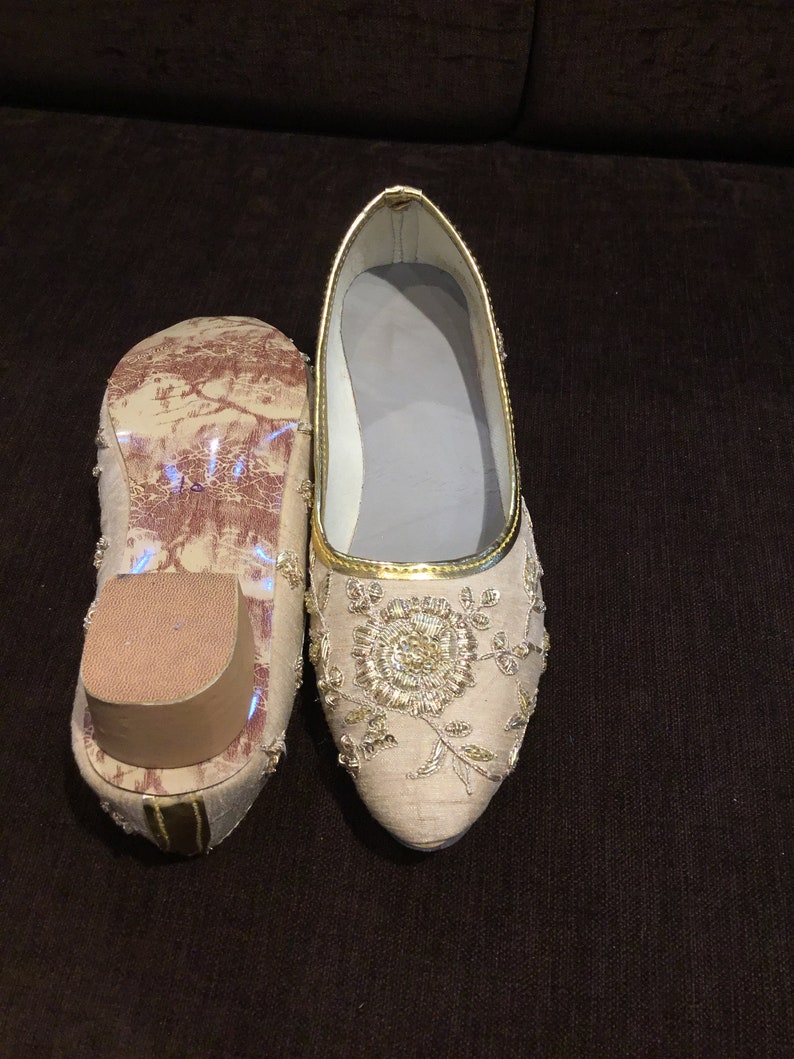 Op de afbeelding: Een paar beige platte schoenen met een puntige neus en gedetailleerd goudkleurig borduurwerk in een bloemmotief. E&eacute;n schoen is van bovenaf zichtbaar, terwijl de andere is omgedraaid om de zool en een kleine blokhak te laten zien.