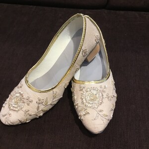 Op de afbeelding: Een paar beige instapschoenen met ingewikkeld goudborduursel en bloemmotieven van lovertjes. De schoenen hebben een puntige neus, een metallic gouden rand langs de opening en een lage blokhak. De binnenvoering is lichtgrijs van kleur.