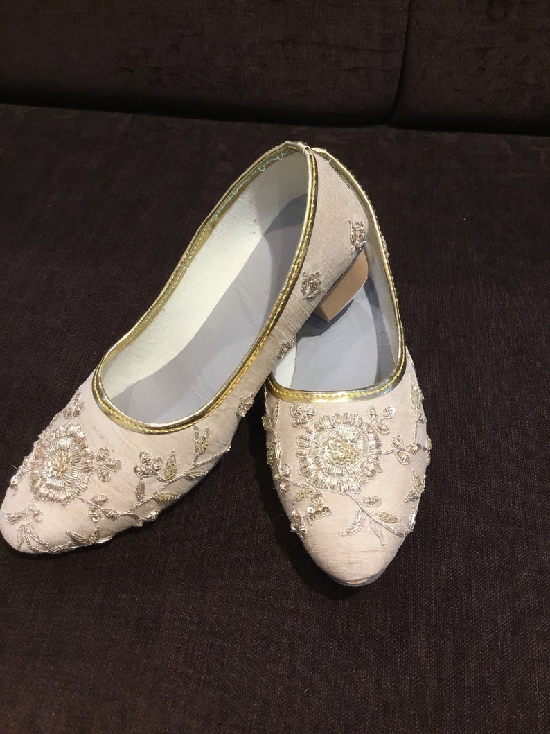 Op de afbeelding: Een paar beige instapschoenen met ingewikkeld goudborduursel en bloemmotieven van lovertjes. De schoenen hebben een puntige neus, een metallic gouden rand langs de opening en een lage blokhak. De binnenvoering is lichtgrijs van kleur.