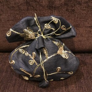 May include: A dark navy blue drawstring pouch featuring intricate gold metallic embroidery in a floral vine pattern. The fabric has a subtle horizontal texture, and the bag is cinched at the top with matching dark navy blue fabric cords.