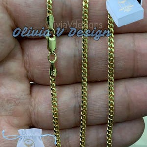 Pode incluir: Um colar de corrente dourada com fecho de lagosta, marcado "925 ITALY". O colar é exibido com um pequeno saco branco com cordão e uma caixa azul clara, ambos com a marca "Olivia V Designs". A imagem é tirada em uma mão.