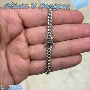 May include: A silver-colored curb chain bracelet with a square clasp, marked "925 ITALY". The bracelet is held in a hand, with a blue gift box and a small blue drawstring bag with the text "Olivia V Designs".