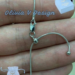 May include: A silver chain necklace with a sliding clasp and two small silver beads. The chain is on a hand and is adjustable in length.