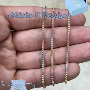 Könnte beinhalten: Drei goldene Ketten, eine silberne, eine roségoldene und eine dreifarbige, werden auf einer Hand gezeigt. Die Ketten sind alle gleich lang und haben ein Gliederketten-Design. Die Ketten befinden sich in einer hellblauen Schachtel mit dem Text "Olivia V. Design" auf der Schachtel.