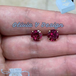 May include: A pair of small, round, red gemstone earrings with gold settings. The earrings are on a person's finger. The earrings are in a small white pouch with the text "Olivia V Design" on it.