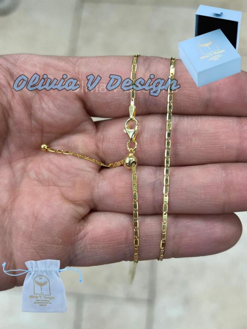 Puede incluir: Un collar de cadena de oro con cierre deslizante. La cadena est&aacute; hecha de peque&ntilde;os eslabones rectangulares. El collar se muestra en una mano y viene con una peque&ntilde;a bolsa blanca y una caja azul. La bolsa y la caja tienen impreso el texto "Olivia V Design".