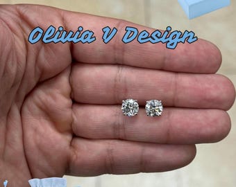 Könnte beinhalten: Ein Paar runde, klare Diamantenohrringe in einer silbernen Fassung. Die Ohrringe werden auf einer Hand gezeigt, im Hintergrund befinden sich eine kleine blaue Tasche und eine blaue Schachtel. Sowohl die Tasche als auch die Schachtel tragen den Aufdruck "Olivia V Design".