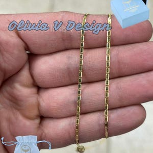 Puede incluir: Un collar de cadena de oro con un dise&ntilde;o de cadena de caja. El collar se muestra en una mano y viene con una peque&ntilde;a caja azul y una bolsa de cord&oacute;n blanco. La caja y la bolsa tienen impreso el texto "Olivia V Design".