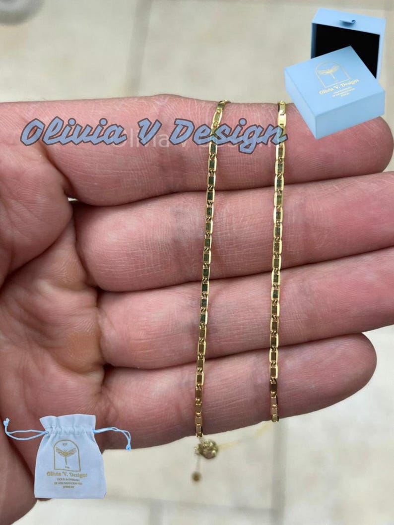 Puede incluir: Un collar de cadena de oro con un dise&ntilde;o de cadena de caja. El collar se muestra en una mano y viene con una peque&ntilde;a caja azul y una bolsa de cord&oacute;n blanco. La caja y la bolsa tienen impreso el texto "Olivia V Design".