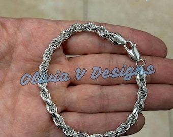 Pode incluir: Uma pulseira de corrente de corda prateada. A pulseira, com elos bem tecidos, possui um fecho seguro. Uma pequena caixa de presente azul e um saco de tecido branco também são visíveis.