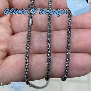 K&ouml;nnte beinhalten: Eine schwarze Kettenkette mit einem Karabinerverschluss. Die Kette besteht aus kleinen, runden Gliedern. Die Kette wird auf einer Hand gezeigt und kommt mit einem kleinen wei&szlig;en Beutel und einer blauen Box.