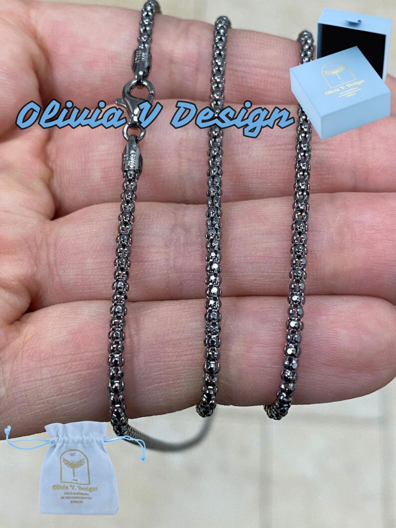 K&ouml;nnte beinhalten: Eine schwarze Kettenkette mit einem Karabinerverschluss. Die Kette besteht aus kleinen, runden Gliedern. Die Kette wird auf einer Hand gezeigt und kommt mit einem kleinen wei&szlig;en Beutel und einer blauen Box.