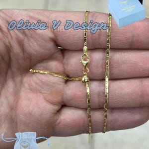 Puede incluir: Un collar de cadena de oro con cierre deslizante. La cadena est&aacute; hecha de peque&ntilde;os eslabones rectangulares. El collar se muestra en una mano, y hay una peque&ntilde;a bolsa blanca y una caja azul en el fondo.