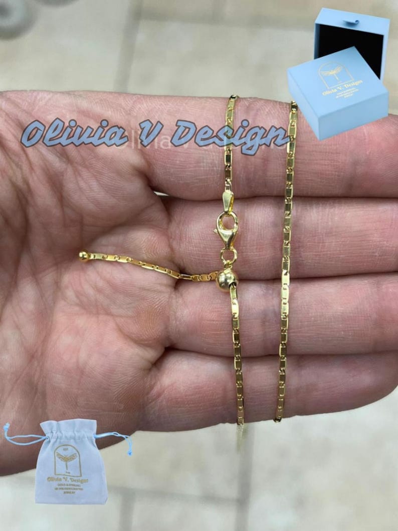 Puede incluir: Un collar de cadena de oro con cierre deslizante. La cadena est&aacute; hecha de peque&ntilde;os eslabones rectangulares. El collar se muestra en una mano, y hay una peque&ntilde;a bolsa blanca y una caja azul en el fondo.