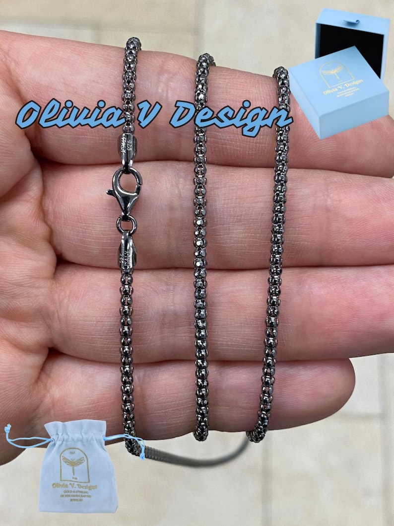 K&ouml;nnte beinhalten: Eine schwarze Kettenkette mit einem Karabinerverschluss. Die Kette besteht aus kleinen, runden Gliedern. Die Kette wird auf einer Hand gezeigt und kommt mit einer kleinen wei&szlig;en Tasche mit dem Text "Olivia V Design" geliefert.