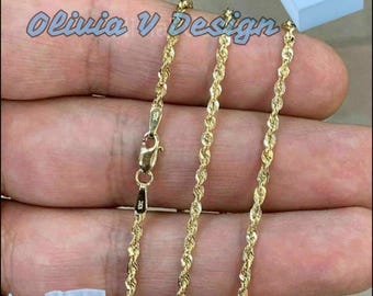 May include: A gold rope chain necklace with a clasp. The necklace is displayed on a hand and comes with a blue box and a white pouch with the text "Olivia V Design".