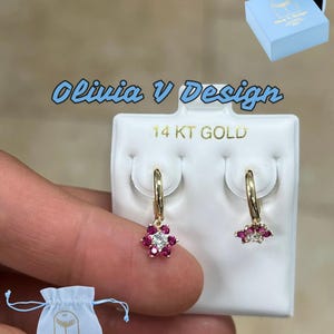 May include: A pair of 14KT gold hoop earrings with a dangling flower charm. The flower charm is made of red and white stones.