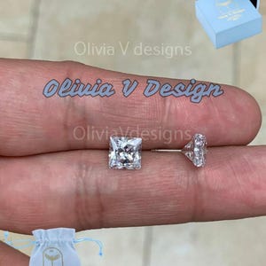 May include: A pair of silver earrings with clear square-shaped cubic zirconia stones. The earrings are displayed on a finger and come with a small blue drawstring bag with the brand name "Olivia V Designs" printed on it.