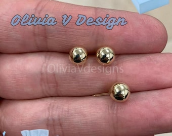 May include: A pair of gold ball stud earrings. The earrings are shown on a hand, with the text "Olivia V Design" visible in the background.