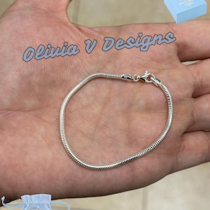 Puede incluir: Una pulsera de cadena de serpiente de color plateado con un cierre de mosquetón. La pulsera se muestra en una mano, con el texto "Olivia V Designs" en azul claro. También se ven una pequeña caja de regalo azul y una bolsa con cordón.