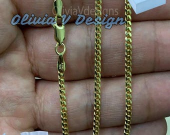 Könnte beinhalten: Eine goldene Kettenkette im Curb-Link-Stil. Die Kette wird auf einer Hand gezeigt und liegt flach. Die Kette befindet sich in einer Schachtel und einem kleinen Beutel mit dem Markennamen "Olivia V Designs" darauf.