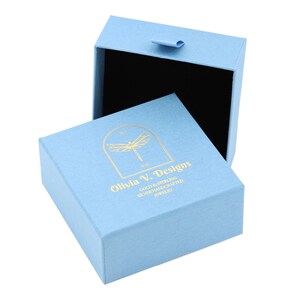 Puede incluir: Una caja de joyer&iacute;a azul claro con un logotipo de lib&eacute;lula dorado y el texto "Olivia V. Designs Gold & Sterling Silver Handcrafted Jewelry".
