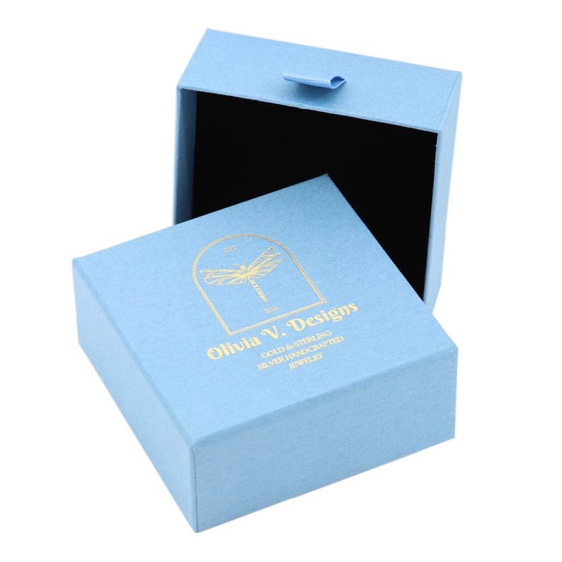 Puede incluir: Una caja de joyer&iacute;a azul claro con un logotipo de lib&eacute;lula dorado y el texto "Olivia V. Designs Gold & Sterling Silver Handcrafted Jewelry".