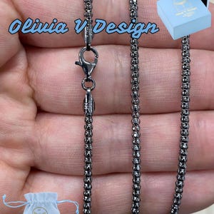 K&ouml;nnte beinhalten: Eine schwarze Kettenkette mit einem Karabinerverschluss. Die Kette besteht aus kleinen, runden Perlen. Die Kette wird auf einer Hand pr&auml;sentiert und kommt mit einem kleinen wei&szlig;en Beutel und einer blauen Box.