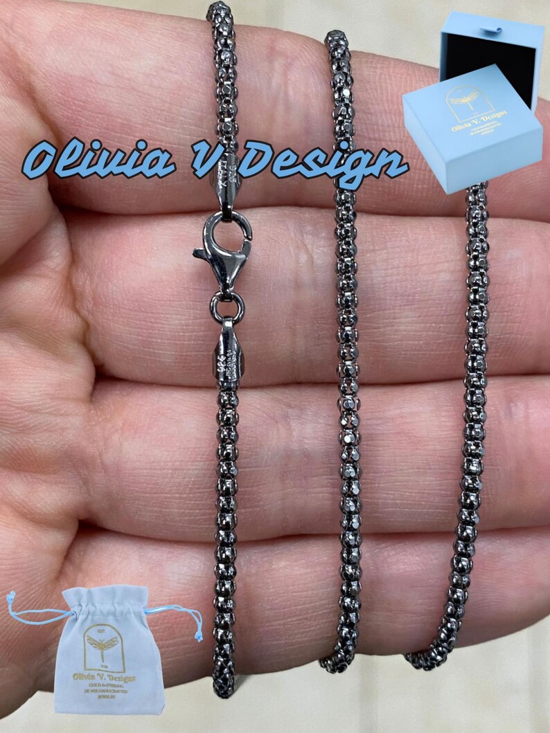 K&ouml;nnte beinhalten: Eine schwarze Kettenkette mit einem Karabinerverschluss. Die Kette besteht aus kleinen, runden Perlen. Die Kette wird auf einer Hand pr&auml;sentiert und kommt mit einem kleinen wei&szlig;en Beutel und einer blauen Box.